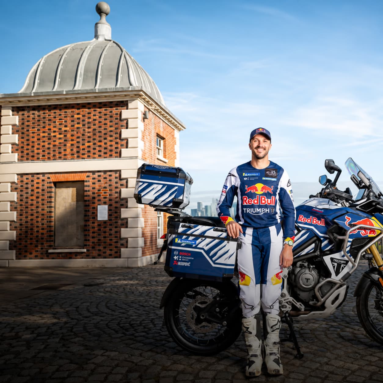 Full-length portrait of professional rider Sam Sunderland wearing Red Bull and Triumph riding kit, standing on a cobblestone courtyard with a fully equipped Triumph Tiger 1200 Rally Explorer adventure bike featuring luggage panniers behind him.