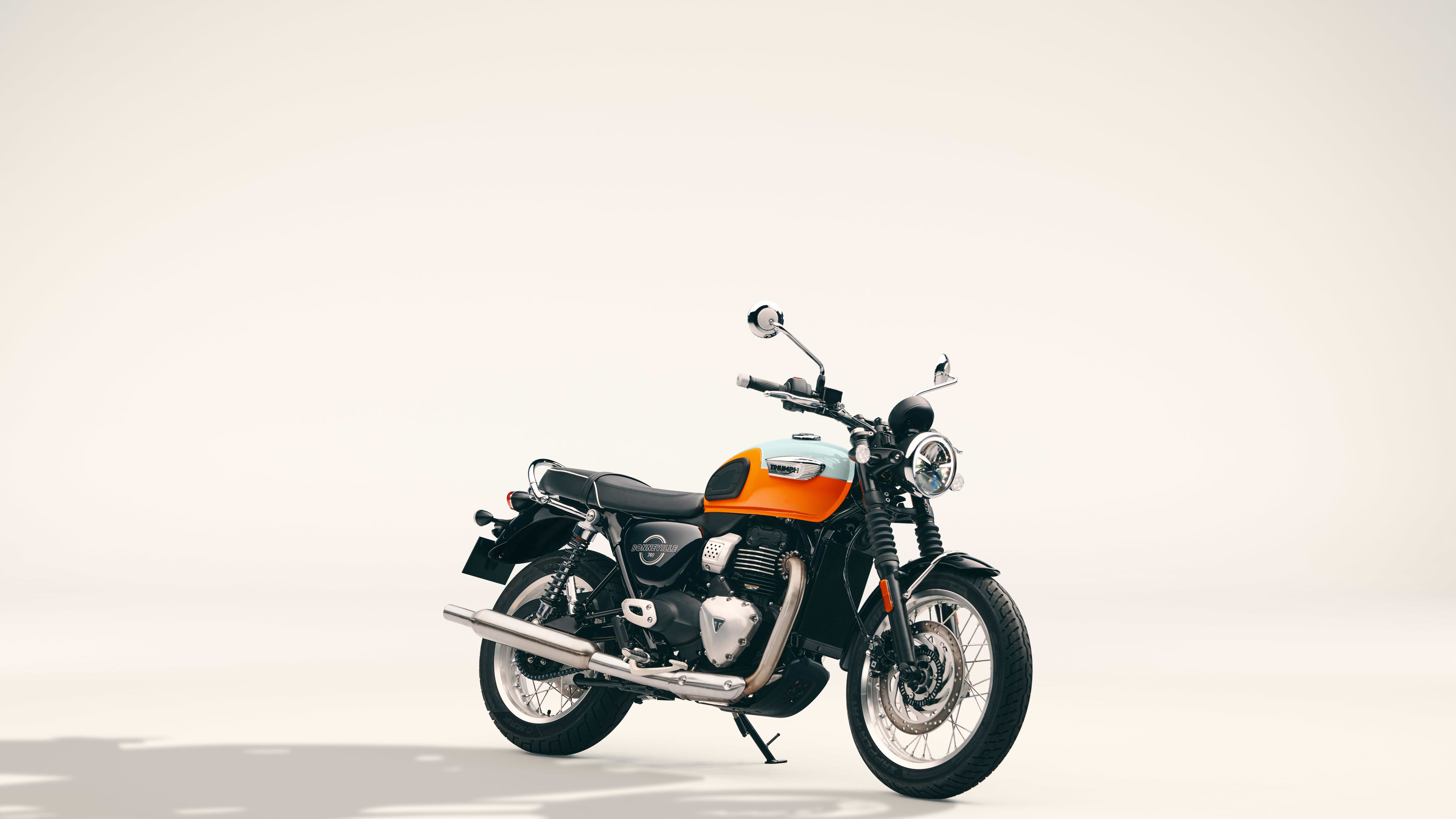 Studio shot of an orange and white Triumph Bonneville T40 motorcycle with chrome exhaust parked on a light background.