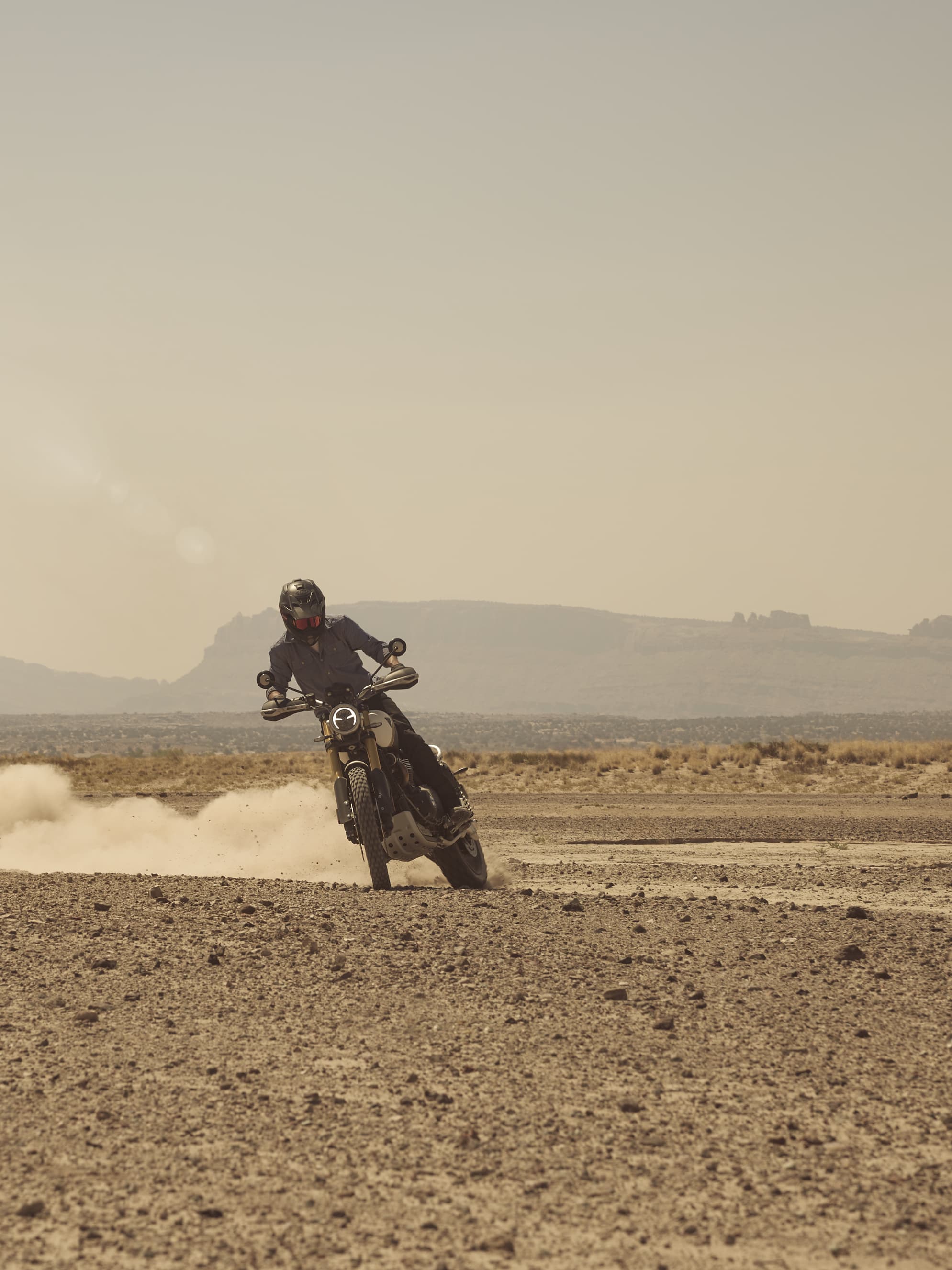 Stil. Ausstrahlung. Funktionalität. Rider powers through rugged terrain on a Triumph Scrambler 1200 XE, kicking up dust against a dramatic outdoor backdrop.