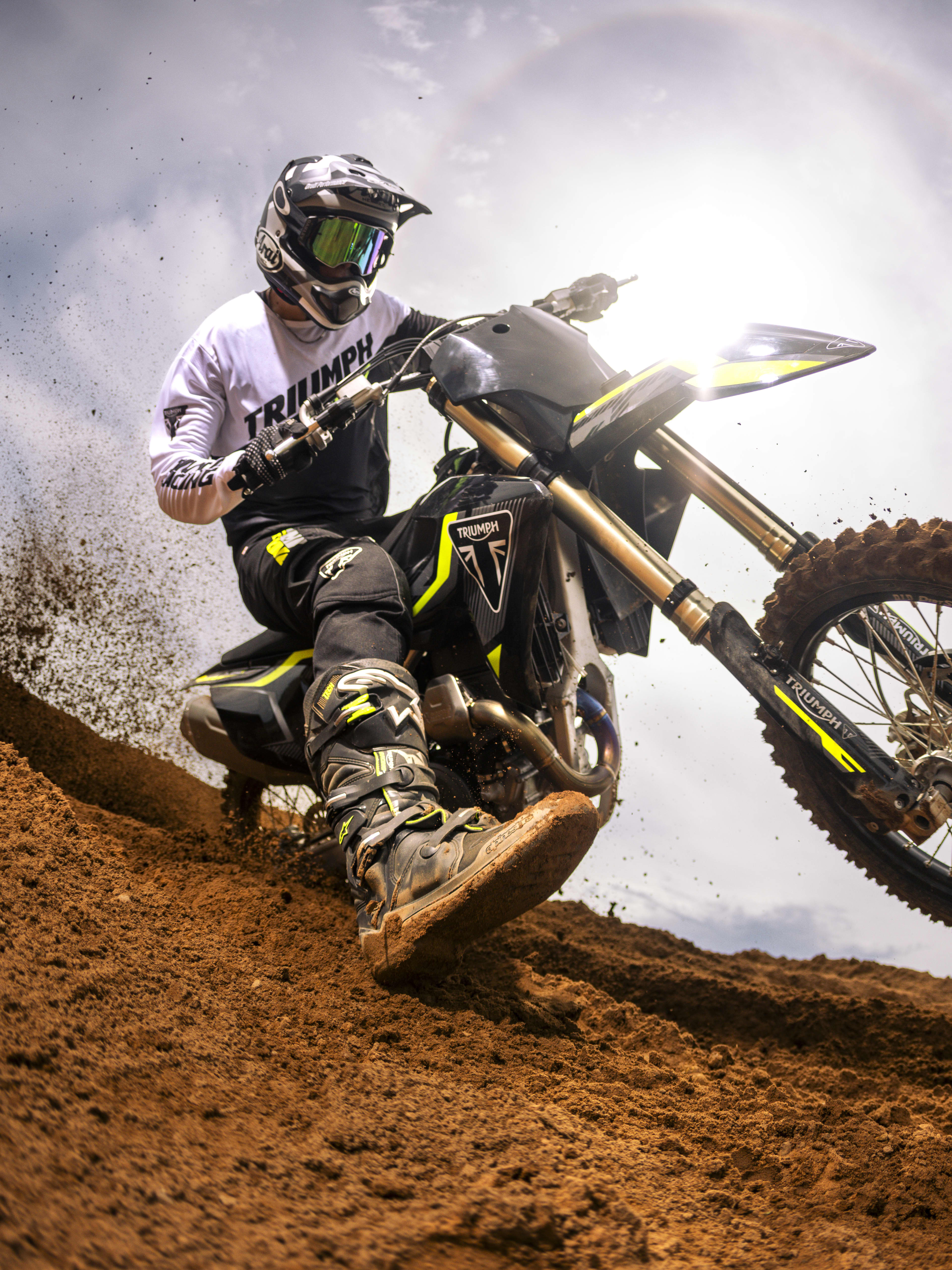 Dramatic low-angle action shot of a helmeted rider in Triumph gear powering up a steep dirt berm on a Triumph TF 250-X motocross bike, highlighting the front KYB suspension, front wheel lifted, and sky backlit by sunlight.