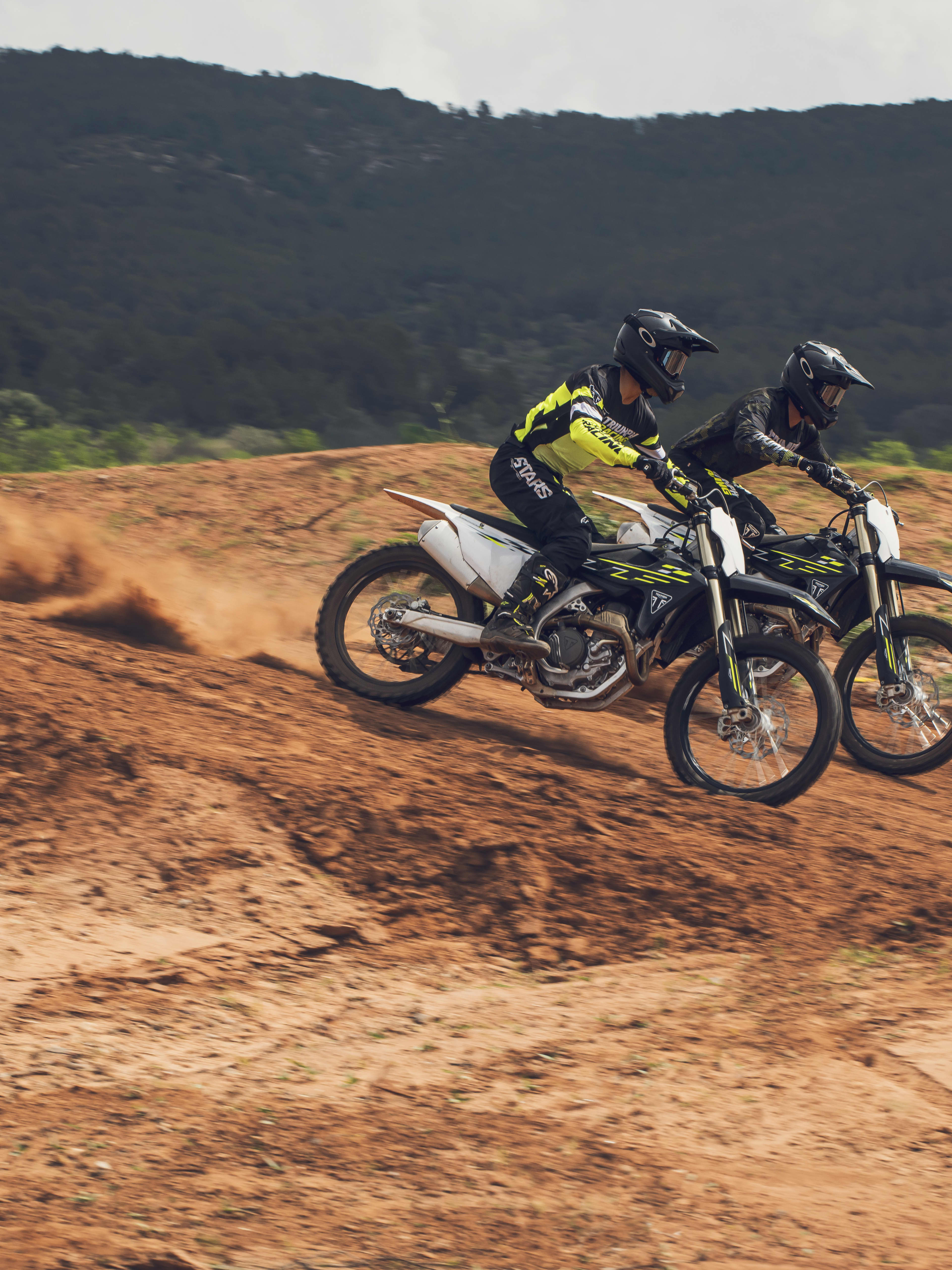 Two off-road motorbike riders in riding gear cresting a red dirt mound side by side on Triumph TF 250-X dirt bikes outdoors.