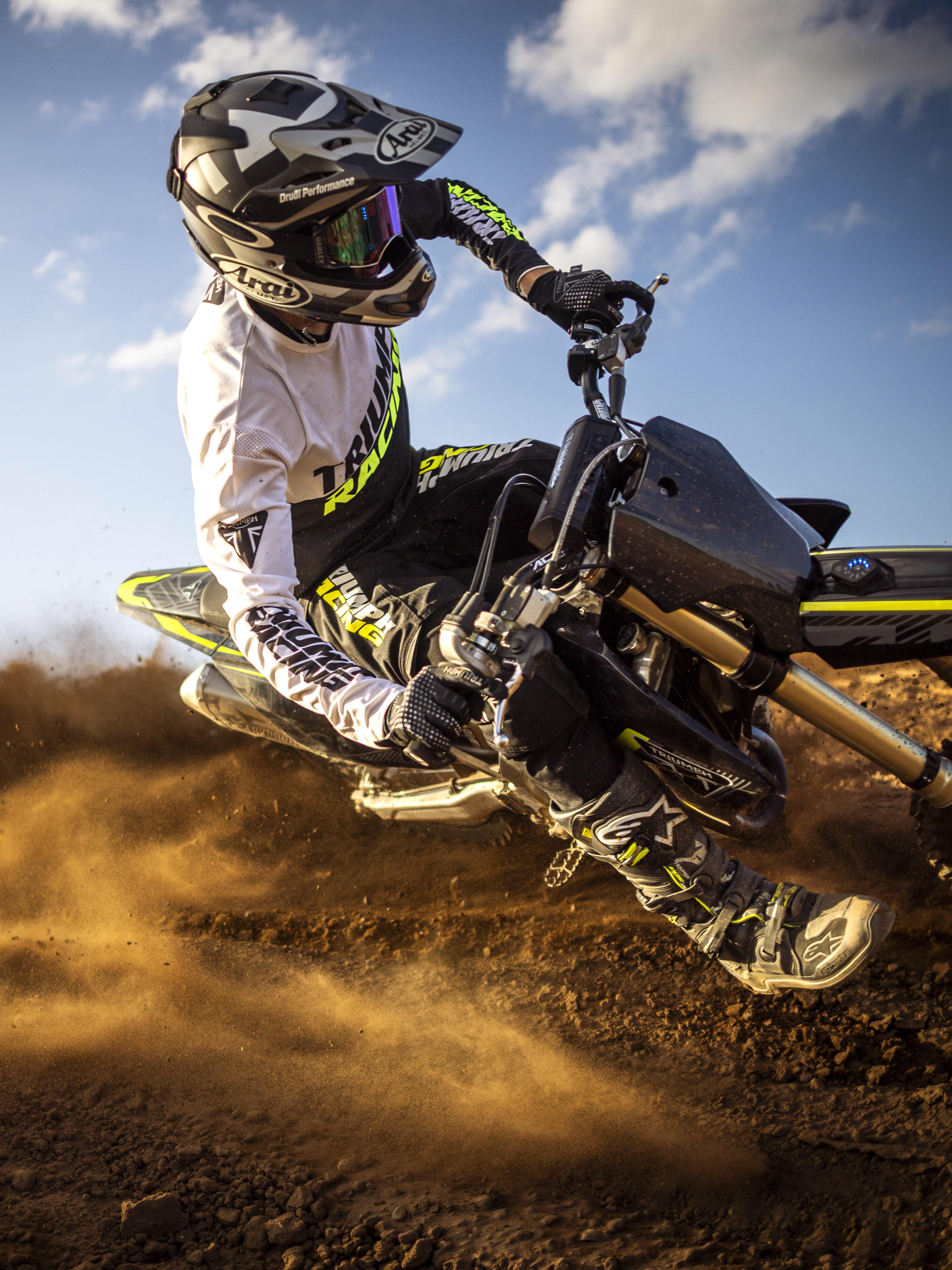 A motocross rider leaning heavily into a turn on a Triumph 250-X motocross bike, kicking up a large cloud of dust.