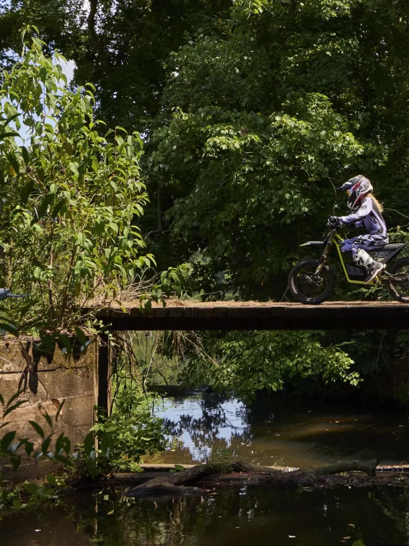 Wide profile shot showing two young riders in full motocross gear crossing a wooden bridge over a creek on Triumph TXP-20 electric dirt bikes, surrounded by dense green forest vegetation.