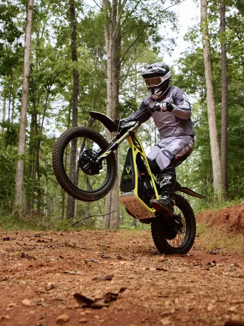 Dynamic wide action shot showing a young rider in full protective gear riding a Triumph TXP-24 electric motorcycle while lifting the front wheel in a wheelie along a red dirt forest trail surrounded by trees.