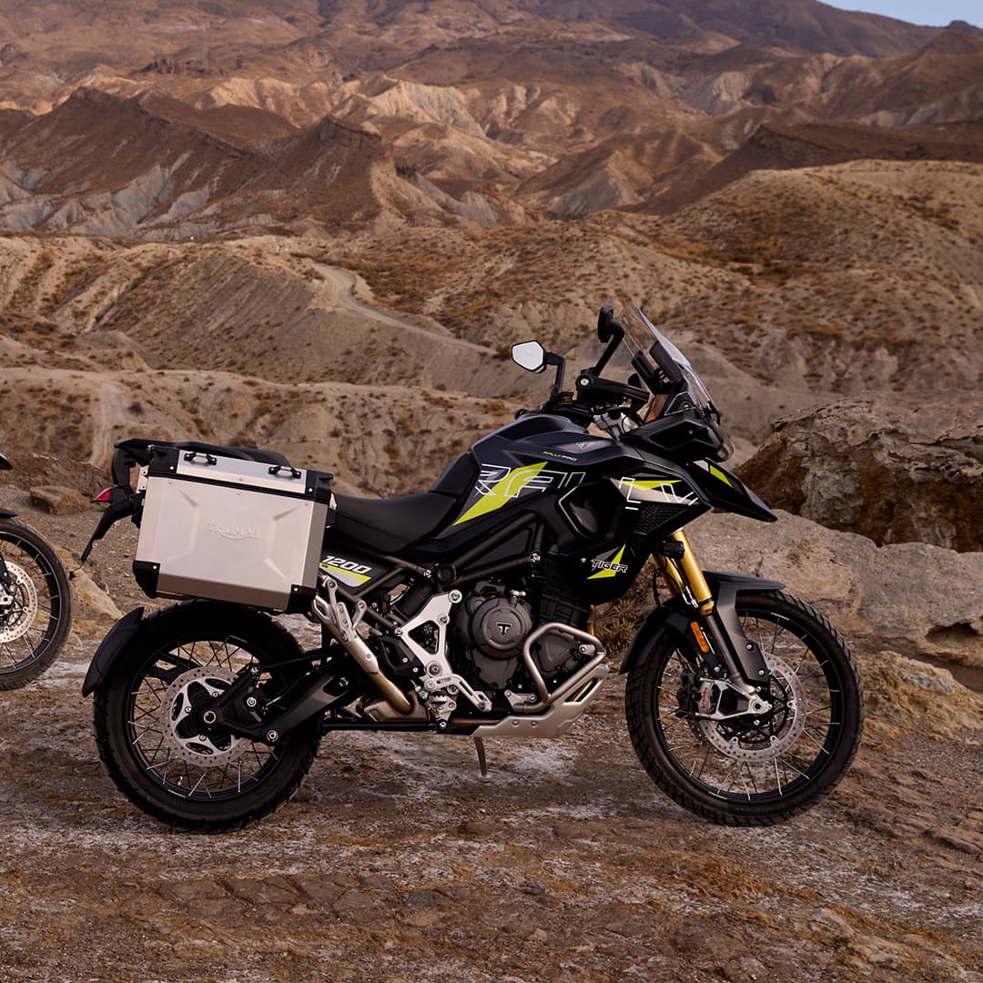 Two Triumph adventure motorcycles—a Tiger 900 and a Tiger 1200—parked on a rocky desert landscape at sunset with mountains in the background.