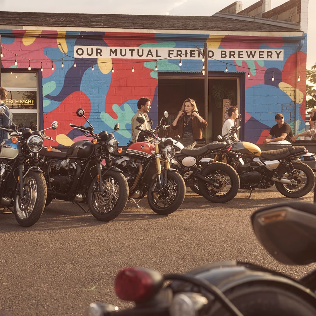 A line of classic Triumph motorcycles parked in front of Our Mutual Friend Brewery, with people socializing in the background under an outdoor mural.