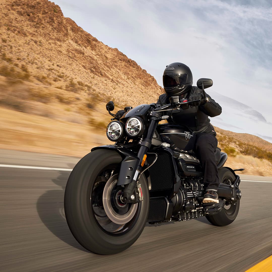 A motorcyclist wearing a black helmet and gear rides a Triumph Rocket 3 Storm GT along a curved desert highway with mountains in the background.