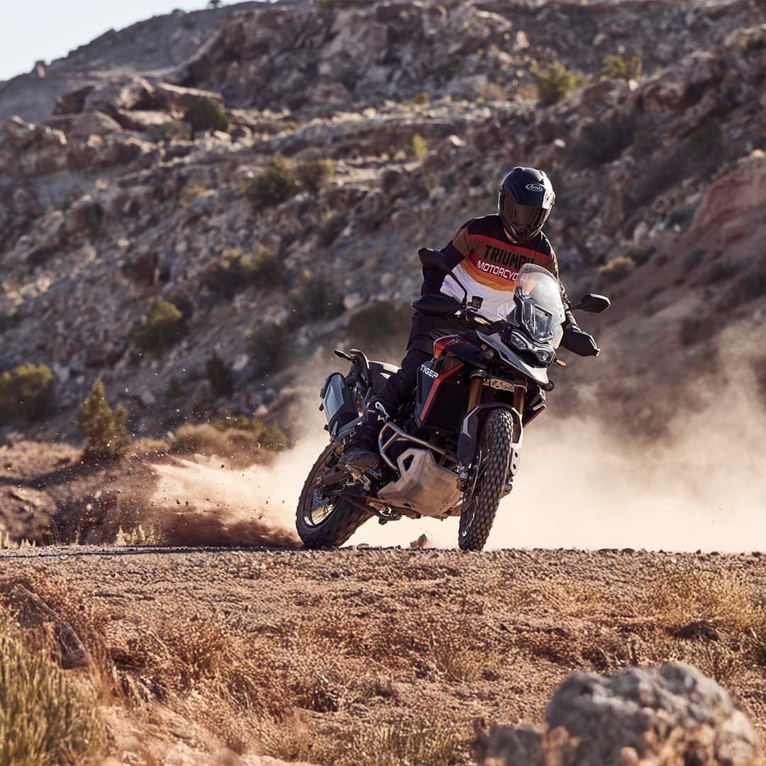 An adventure motorcyclist kicks up dust while riding a Triumph Tiger 900 Rally Pro on an off-road, rugged desert trail.