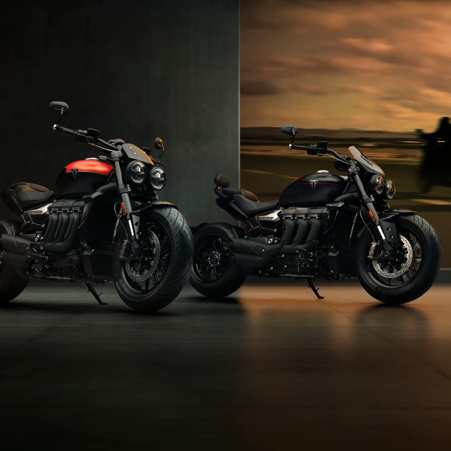 Two Triumph Rocket 3 Storm motorcycles—the R model with a red tank accent and the GT model in all-black—parked side-by-side in a dimly lit studio showroom.