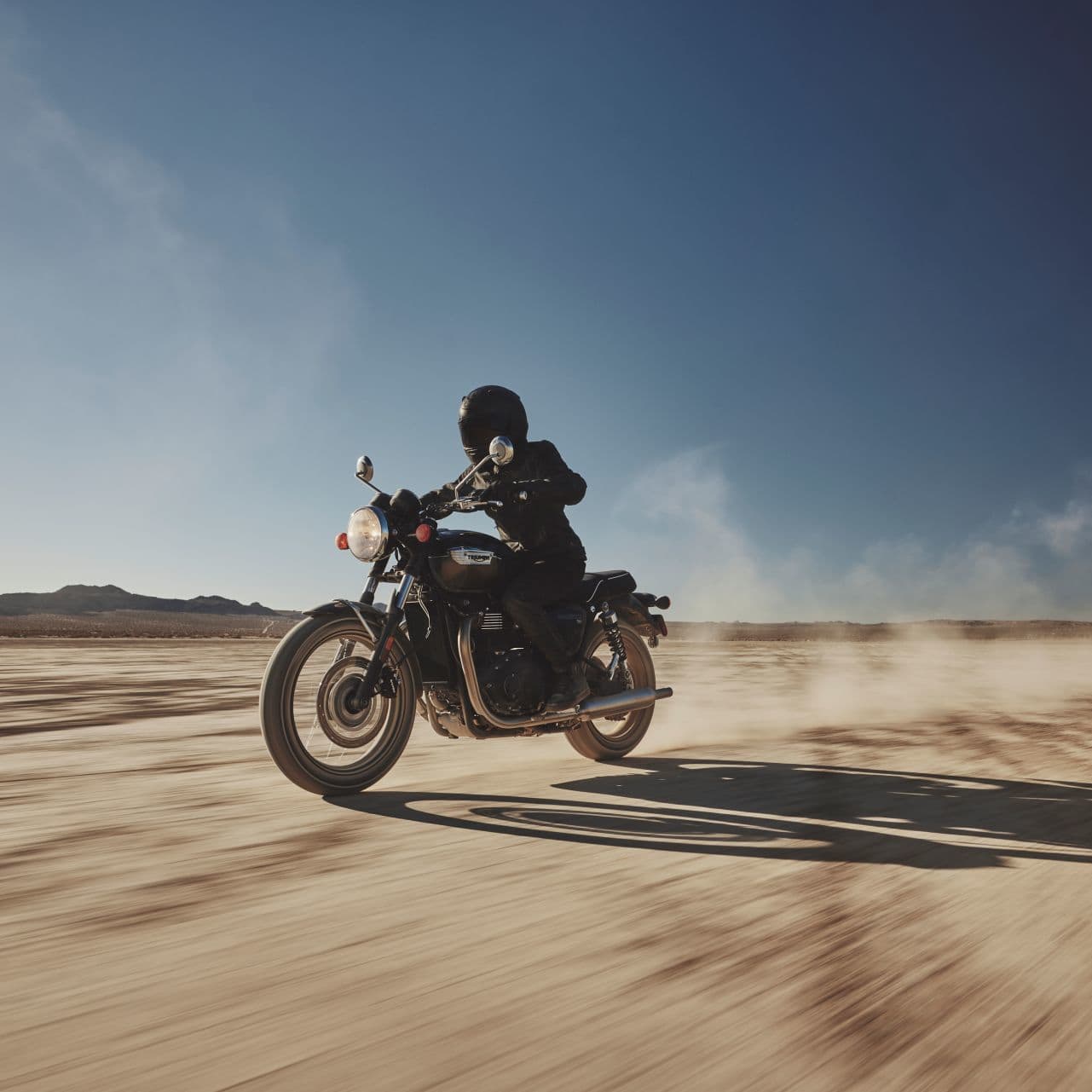 A motorcyclist riding a black Triumph Bonneville T100 across a wide, sunlit desert landscape, kicking up a trail of dust.