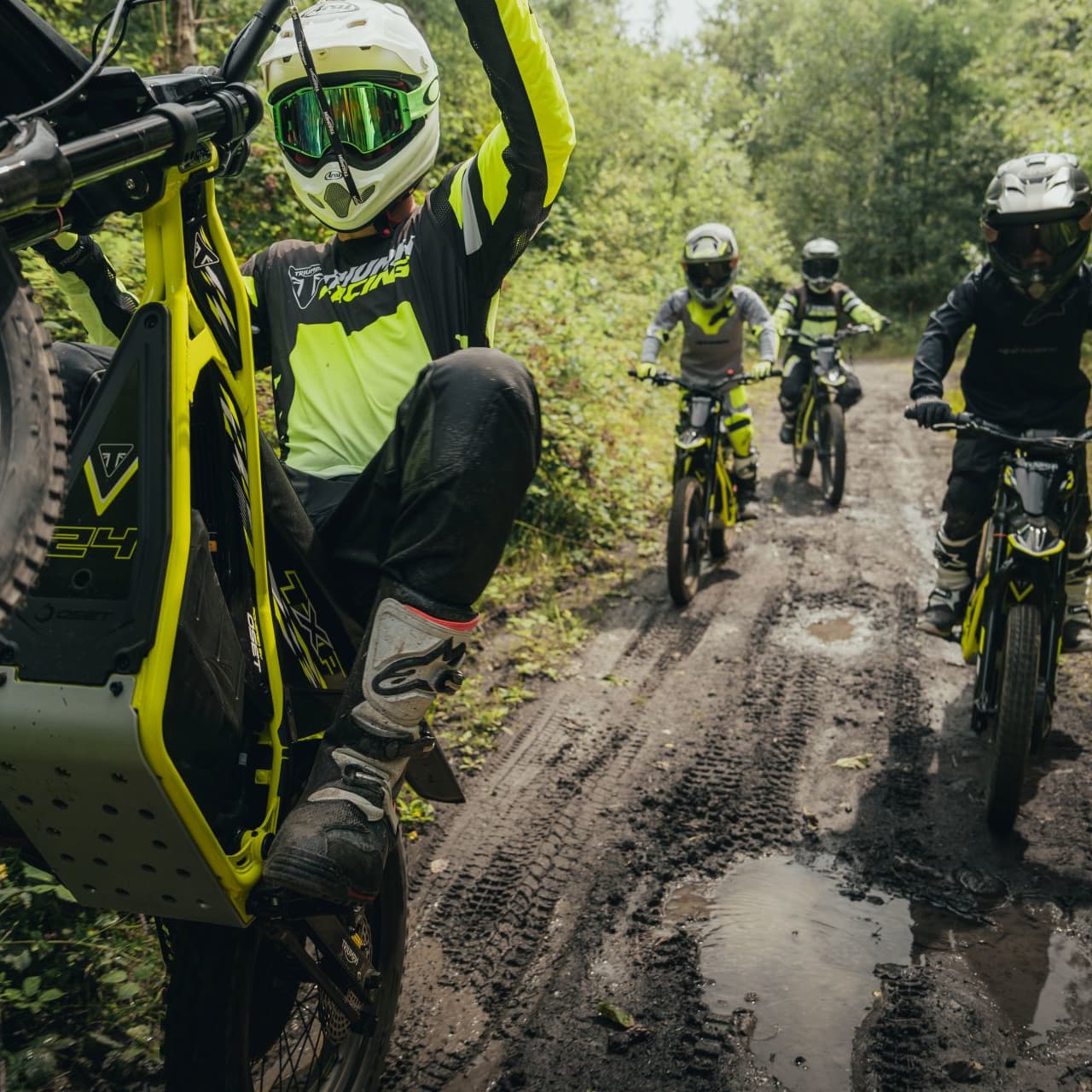 A group of young riders navigating a muddy, tree-lined trail on neon yellow Triumph TXP electric dirt bikes, with the lead rider performing a wheelie.