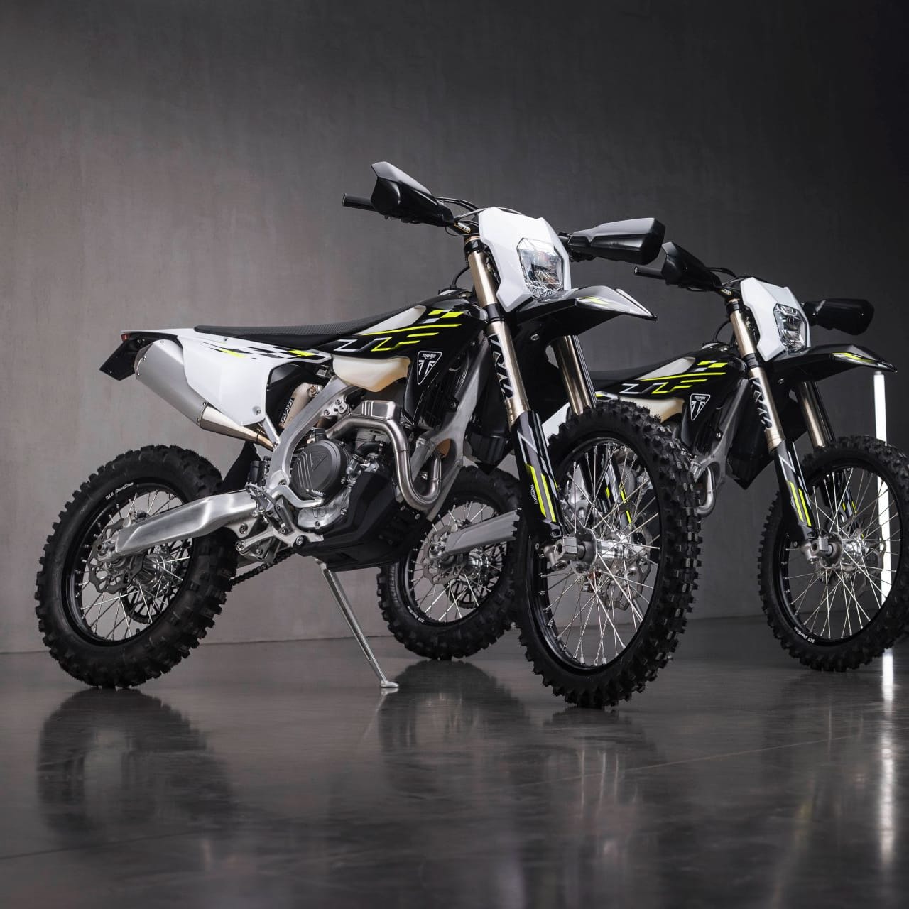Two Triumph enduro motorbikes parked on a glossy dark floor in a studio setting with vertical tube lights.