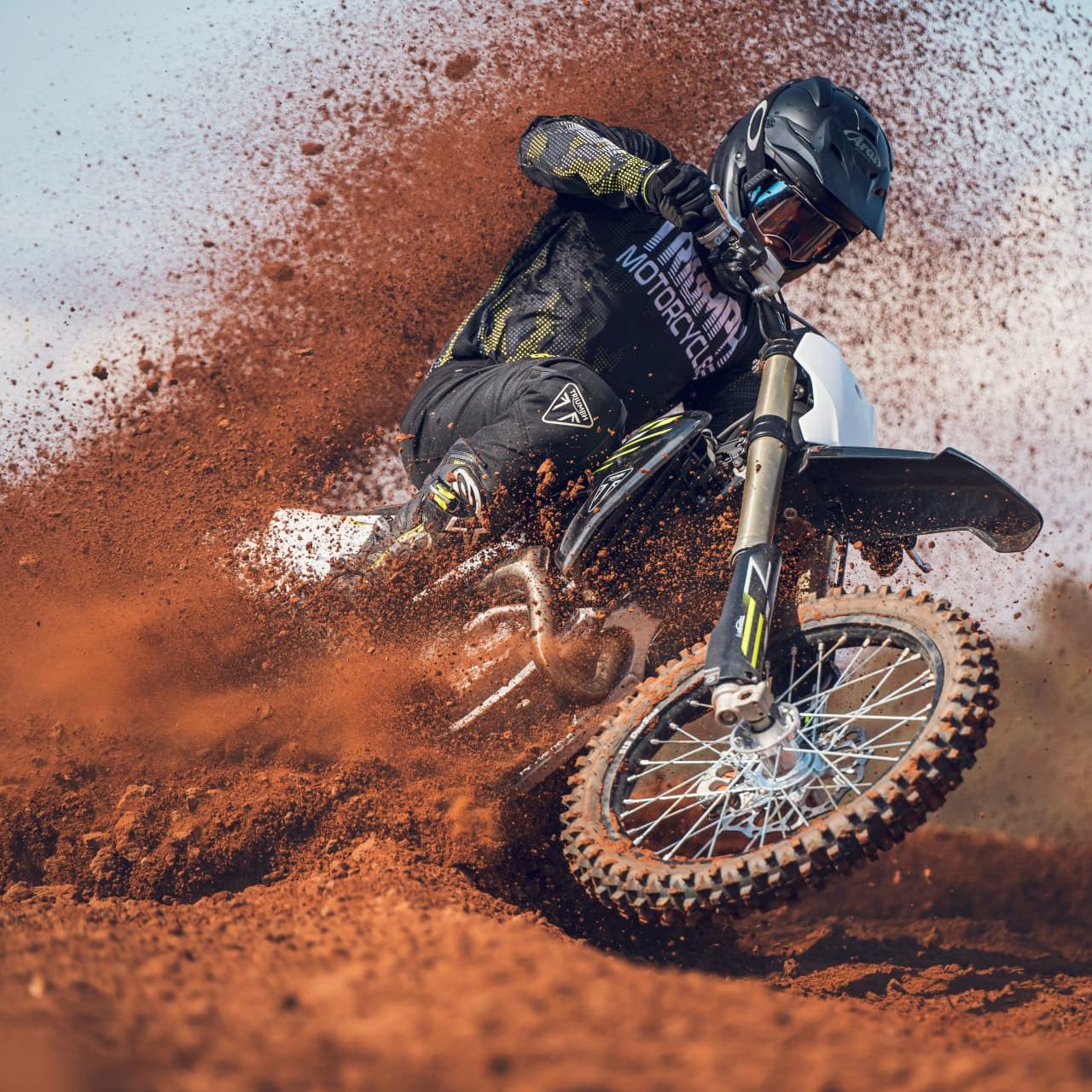A motocross rider in full gear tightly corners a Triumph TF 450-X dirt bike, aggressively blasting thick red dirt and roost into the air.