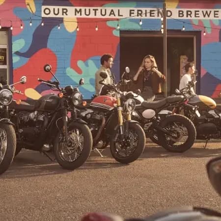 A line of classic Triumph motorcycles parked in front of Our Mutual Friend Brewery, with people socializing in the background under an outdoor mural.