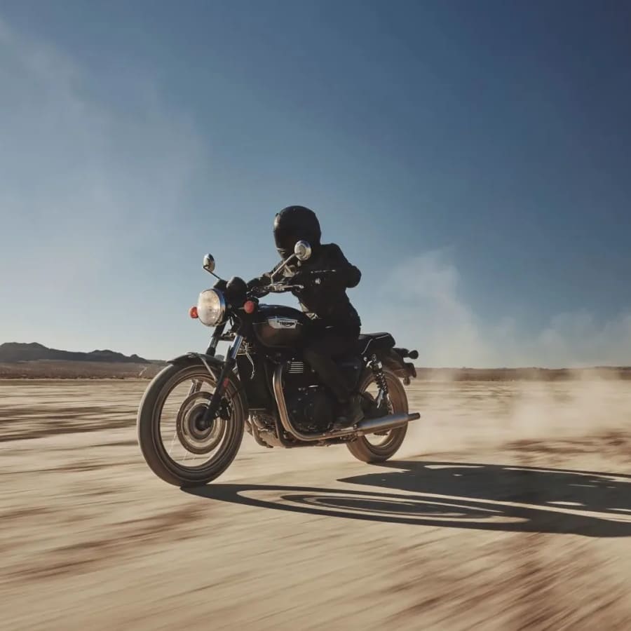 A motorcyclist riding a black Triumph Bonneville T100 across a wide, sunlit desert landscape, kicking up a trail of dust.