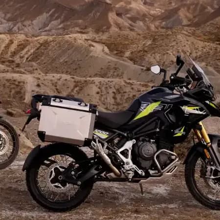 Two Triumph adventure motorcycles—a Tiger 900 and a Tiger 1200—parked on a rocky desert landscape at sunset with mountains in the background.