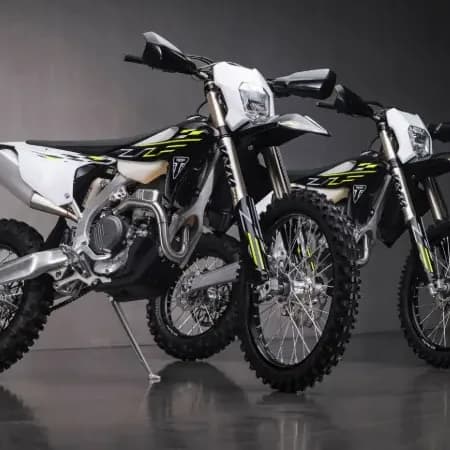 Two Triumph enduro motorbikes parked on a glossy dark floor in a studio setting with vertical tube lights.