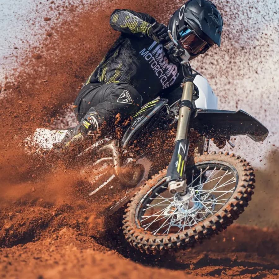 A motocross rider in full gear tightly corners a Triumph TF 450-X dirt bike, aggressively blasting thick red dirt and roost into the air.