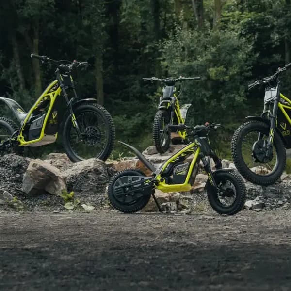 A lineup of four Triumph TXP electric dirt bikes in varying sizes, featuring bright neon yellow frames and knobby tires, parked on a rocky trail with a dense forest background.