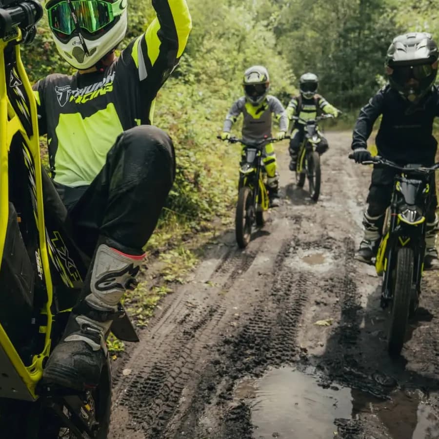 A group of young riders navigating a muddy, tree-lined trail on neon yellow Triumph TXP electric dirt bikes, with the lead rider performing a wheelie.