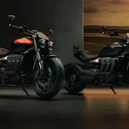 Two Triumph Rocket 3 Storm motorcycles—the R model with a red tank accent and the GT model in all-black—parked side-by-side in a dimly lit studio showroom.