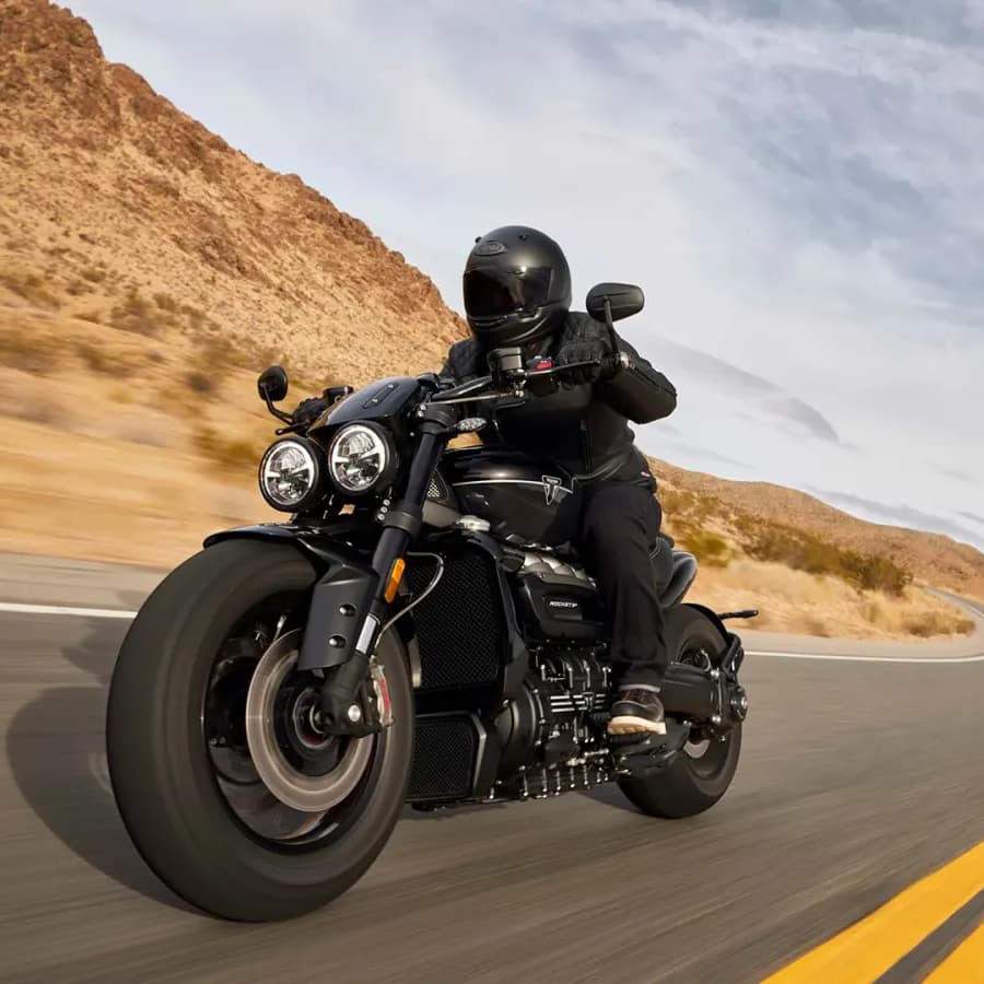 A motorcyclist wearing a black helmet and gear rides a Triumph Rocket 3 Storm GT along a curved desert highway with mountains in the background.