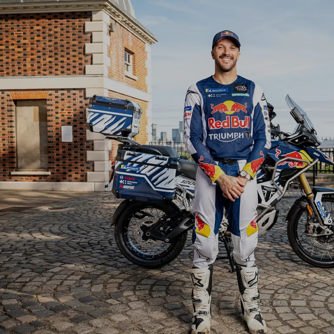 Full-length portrait of professional rider Sam Sunderland wearing Red Bull and Triumph riding kit, standing on a cobblestone courtyard with a fully equipped Triumph Tiger 1200 Rally Explorer adventure bike featuring luggage panniers behind him.